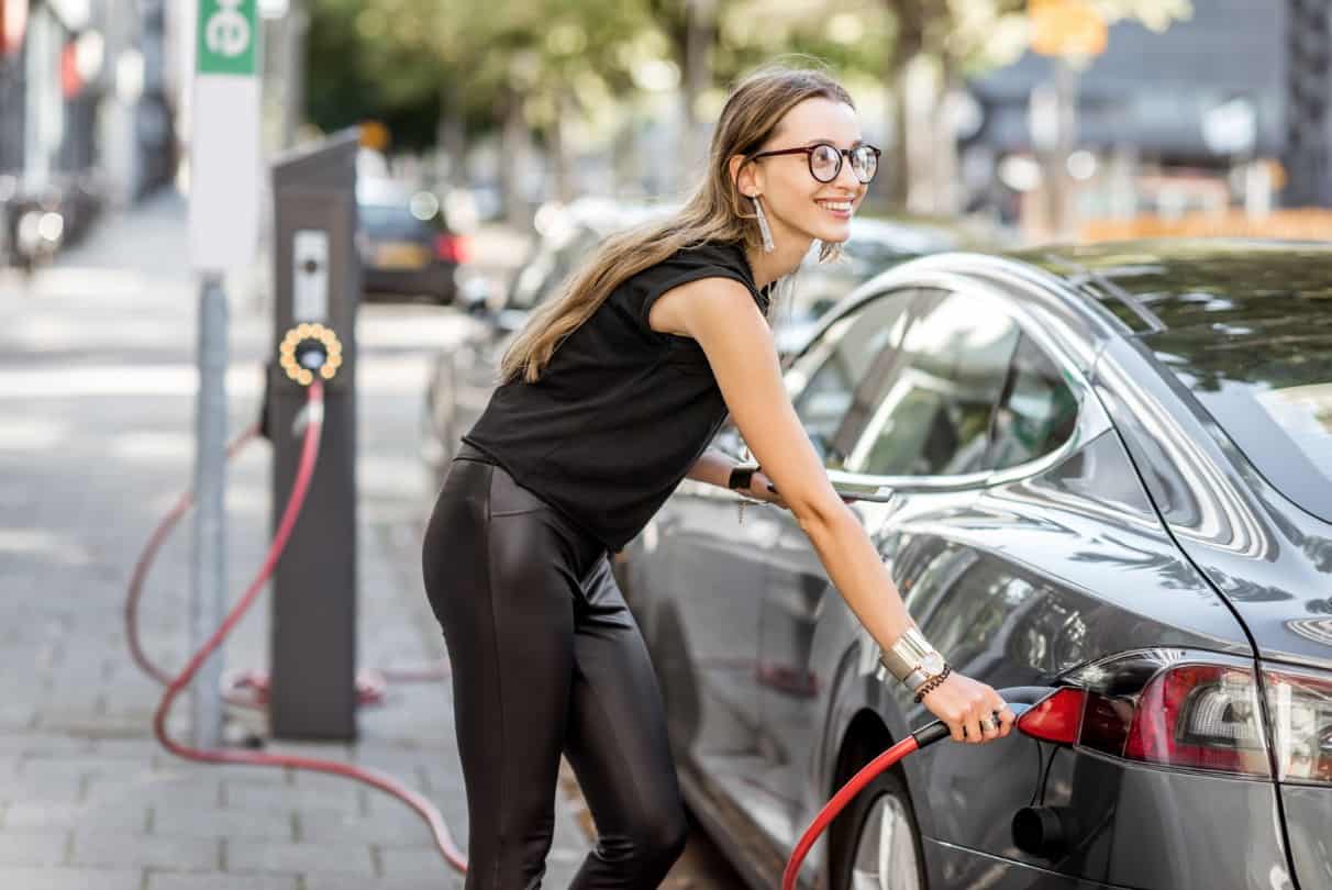 smiling young brunette woman in black charging an electric car